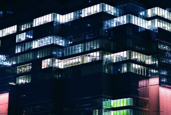 Bâtiment de bureaux moderne la nuit, avec des fenêtres éclairées révélant des intérieurs. Façade sombre aux formes géométriques, lumières rouges et vertes.