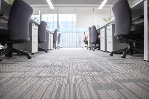 Bureau moderne vide avec moquette grise à motifs. Rangées de bureaux, chaises de bureau sombres et classeurs. Grandes fenêtres lumineuses.