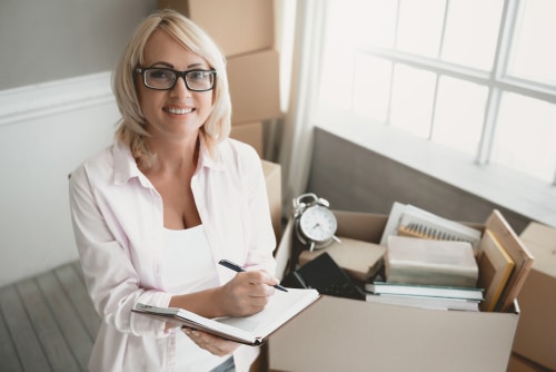 Femme blonde souriante à lunettes, notant sur un carnet près de cartons de déménagement. Elle gère un inventaire.
