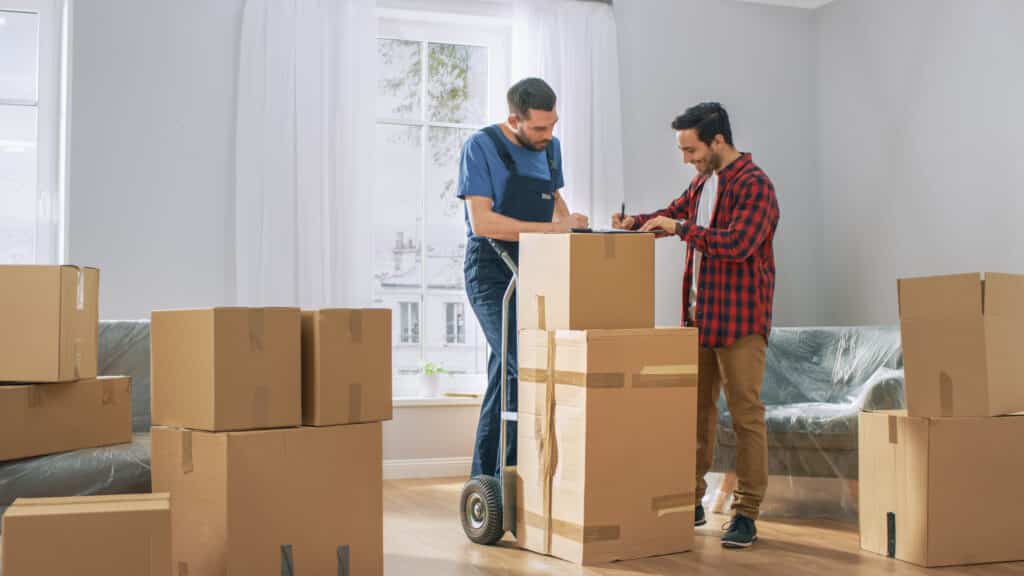 Deux hommes finalisent un déménagement, l'un en uniforme de déménageur présentant des documents à l'autre souriant, près de cartons.