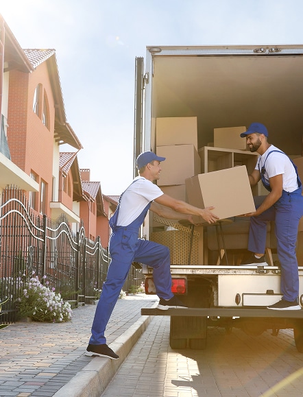 Deux déménageurs souriants en uniforme bleu chargent des boîtes en carton dans un camion de déménagement, devant des maisons.