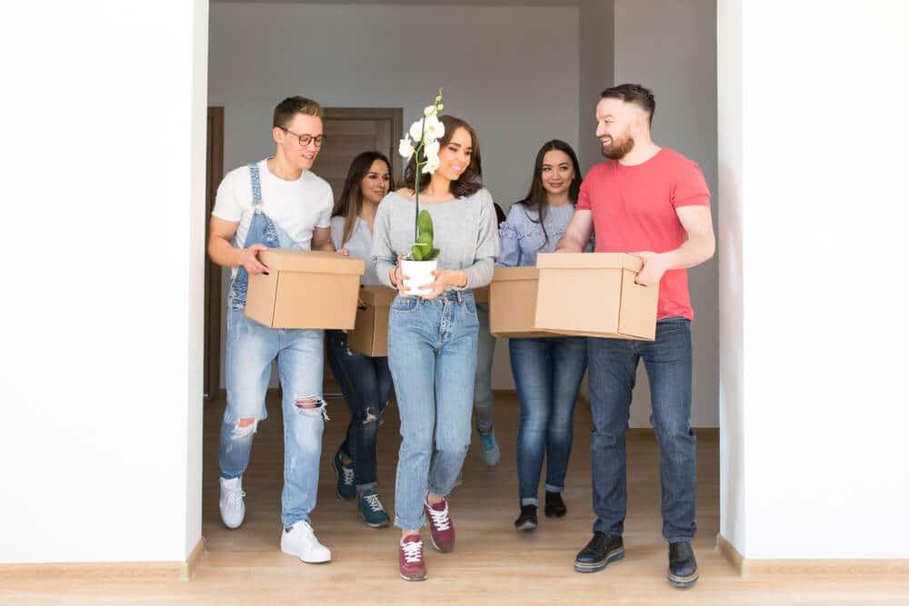 Six amis souriants, dont un homme en salopette et une femme avec une orchidée, emménagent en portant des cartons.