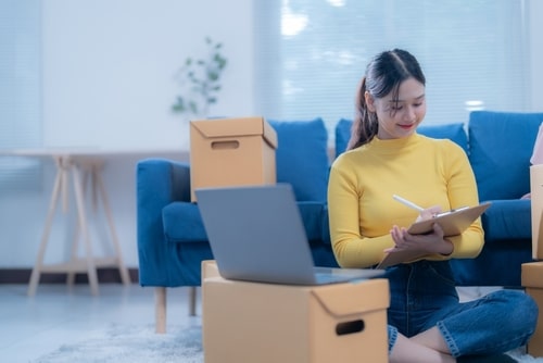 Jeune femme souriante en pull jaune, écrivant sur un presse-papiers près d'un ordinateur portable. Des cartons d'expédition l'entourent, suggérant un business en ligne.