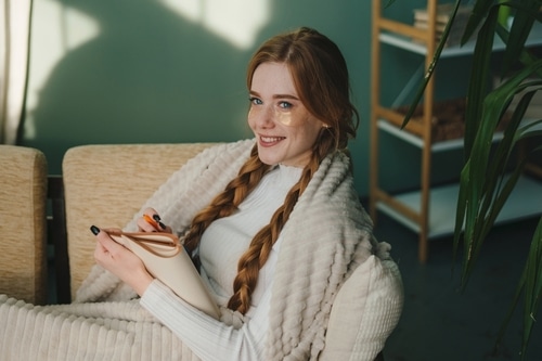 Femme rousse souriante avec patchs yeux dorés, enveloppée dans une couverture douce, tenant un carnet et un stylo orange.