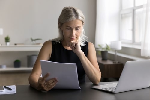 Femme blonde concentrée lisant des documents, main au menton, devant un ordinateur portable sur un bureau moderne.