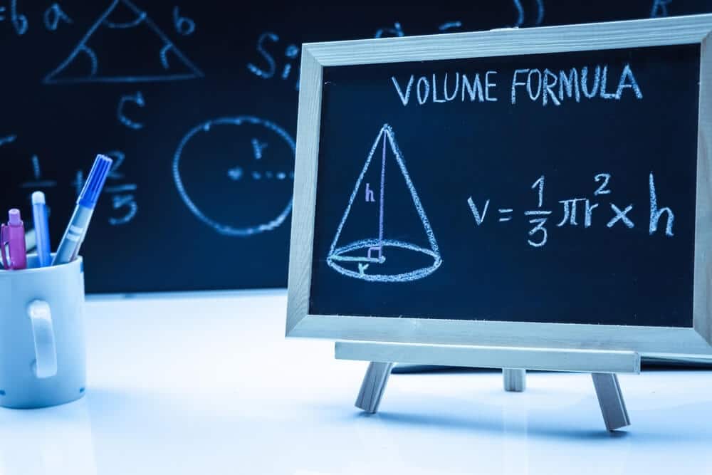 Tableau noir miniature "VOLUME FORMULA" et dessin d'un cône avec V=1/3πr²h. Mug de stylos et tableau de maths en arrière-plan.