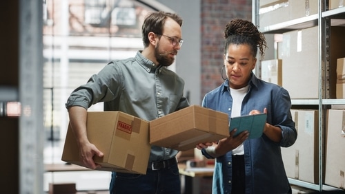 Homme et femme gérant la logistique en entrepôt. L'homme tient des colis dont un "FRAGILE", la femme vérifie l'inventaire sur tablette.