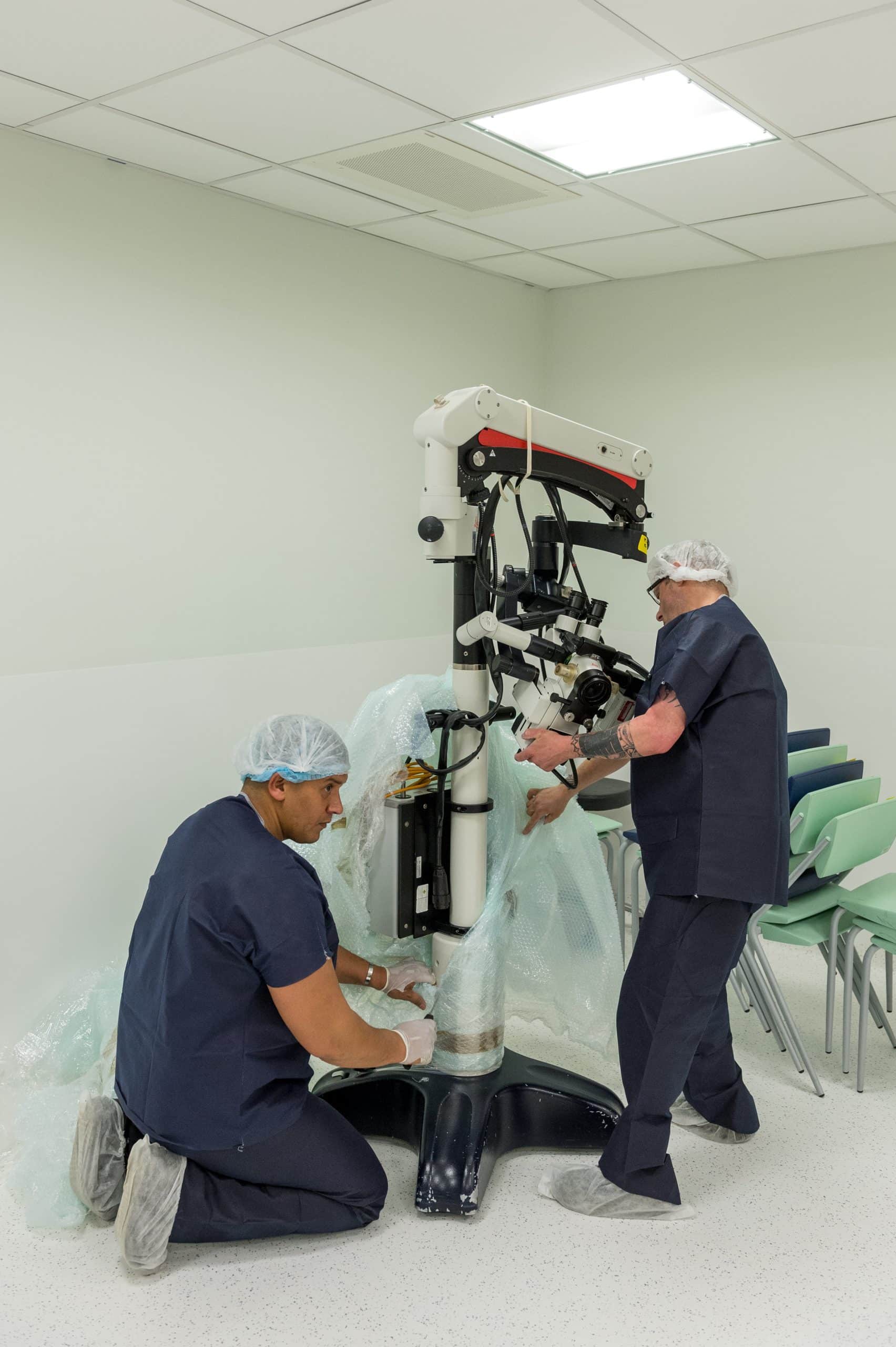 Deux professionnels en tenue médicale, charlotte et sur-chaussures, installent un grand microscope chirurgical dans une salle d'opération lumineuse.