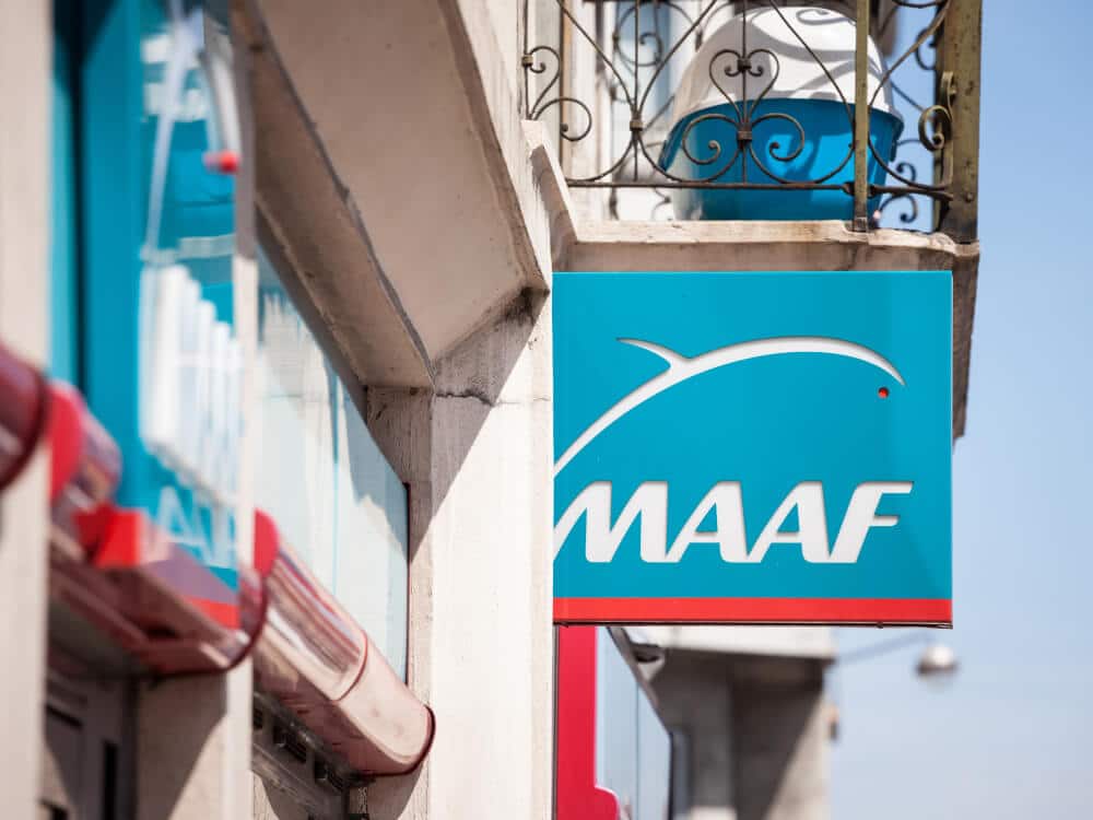 Enseigne bleue de l'agence MAAF avec logo et texte blanc, fixée à un bâtiment en pierre claire sous ciel bleu. Gouttières rouges visibles.