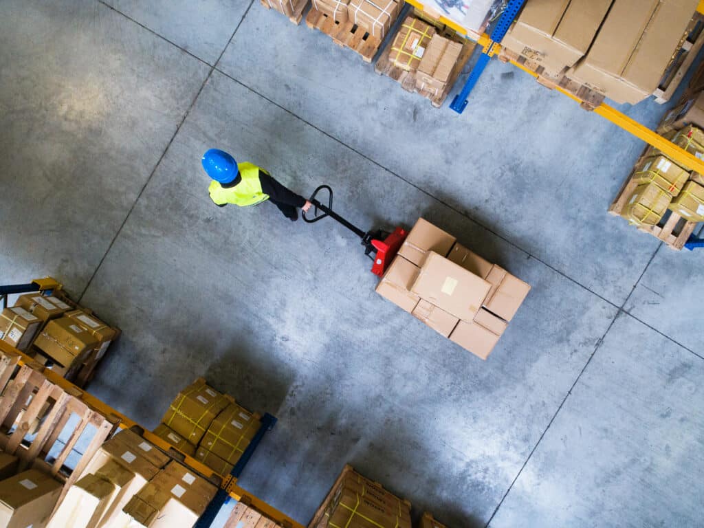 Vue aérienne d'un travailleur en gilet jaune et casque bleu poussant un transpalette rouge chargé de cartons dans un entrepôt. Rayonnages remplis de marchandises.