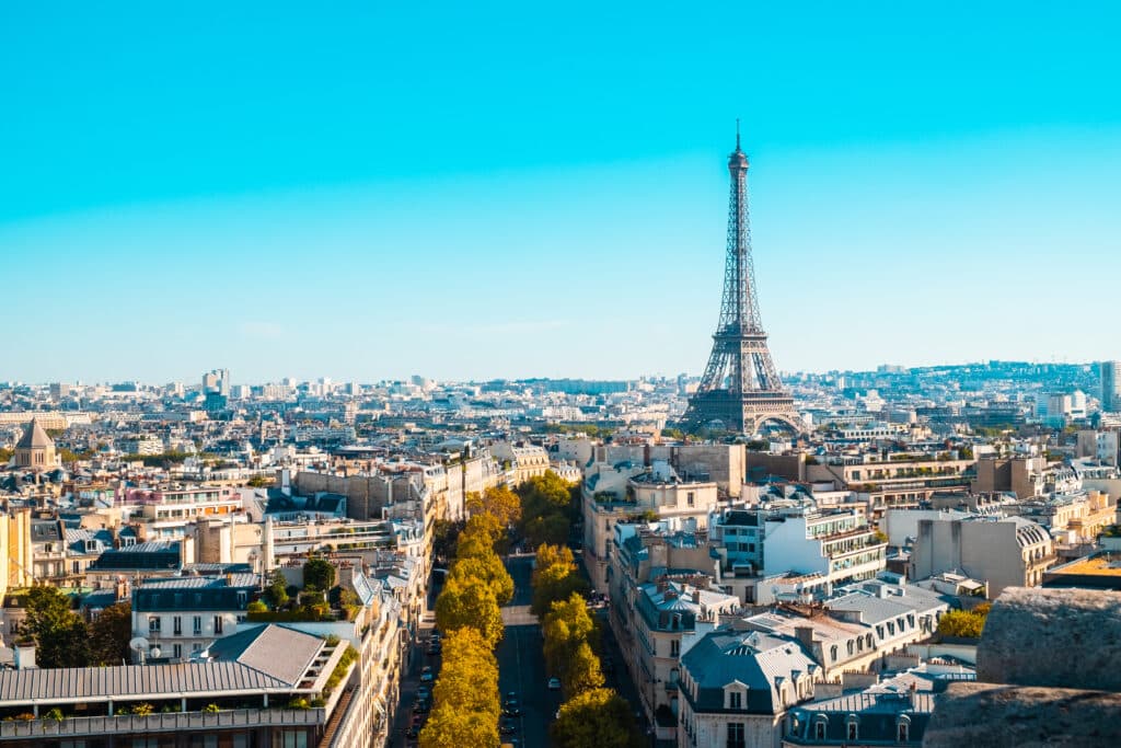 Vue panoramique de Paris avec la Tour Eiffel sous un ciel bleu. Toits haussmanniens et avenue bordée d'arbres au premier plan.