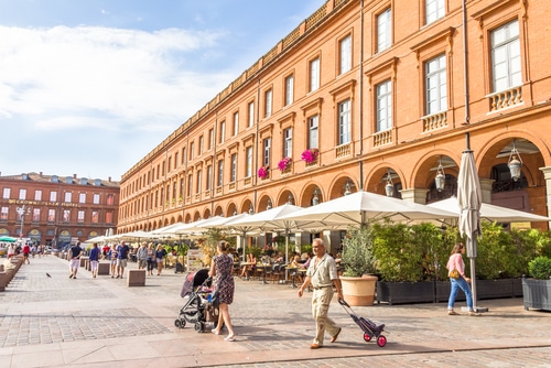 Place de Toulouse, bâtiments en brique orange, arcades abritant des terrasses de café animées sous parasols. Gens se promenant, Midoway Plaza Hotel.